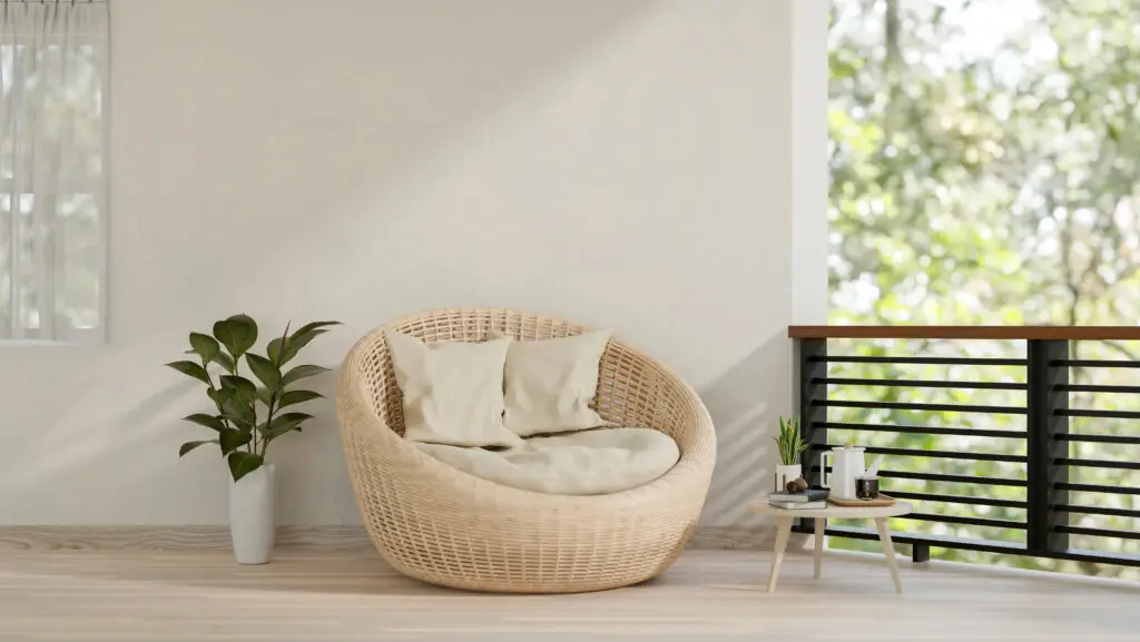 Ronde rotan stoel met kussens en een plant in een lichte ruimte met uitzicht op groen.